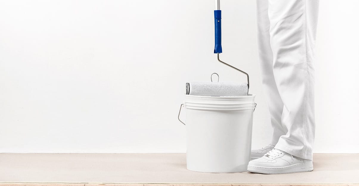 un seau de peinture avec un rouleau et le bas des jambes d’un peintre